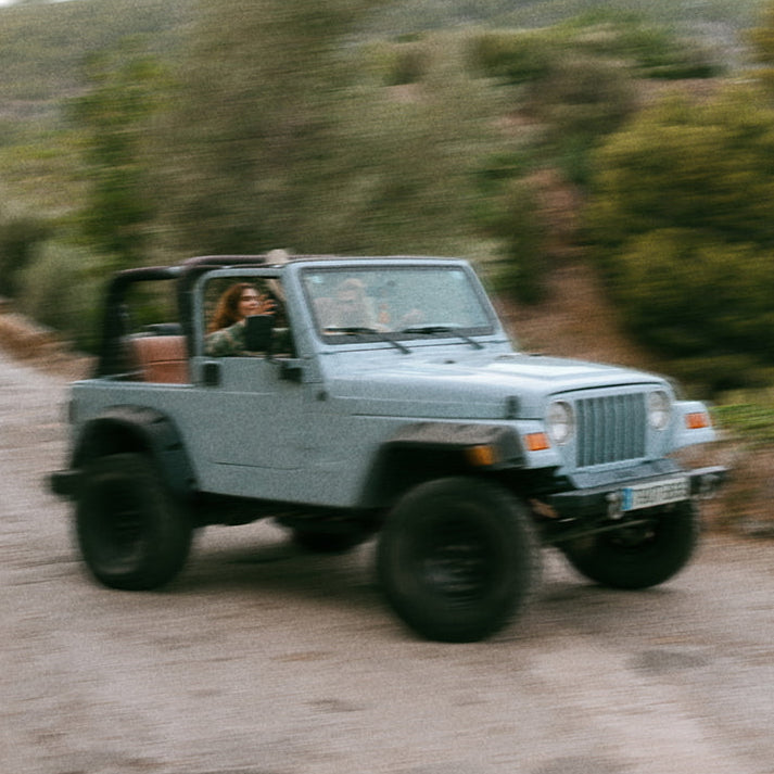 A light blue vintage Jeep Wrangler driving on a dirt road with blurred trees and hills in the background, showing a person inside the vehicle.